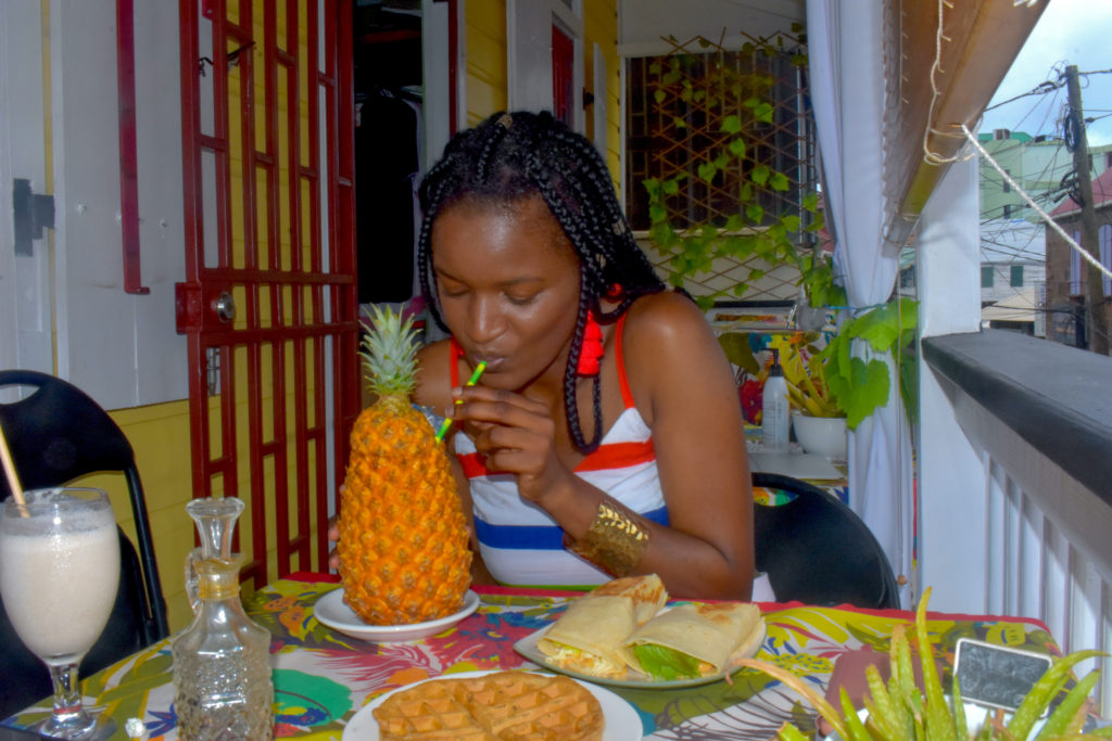 Black african girl sipping a pina colada with healthy island food in the caribbean, wearing red, blue, green and white african overalls at a tropical island cafe