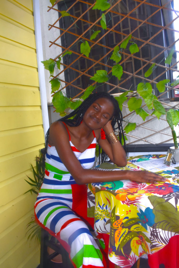 Black african girl enjoying the day waiting for healthy island food at Caribbean restaurant and cafe, wearing colorful african print, ankara top and bottom sets and overalls, with box braids.