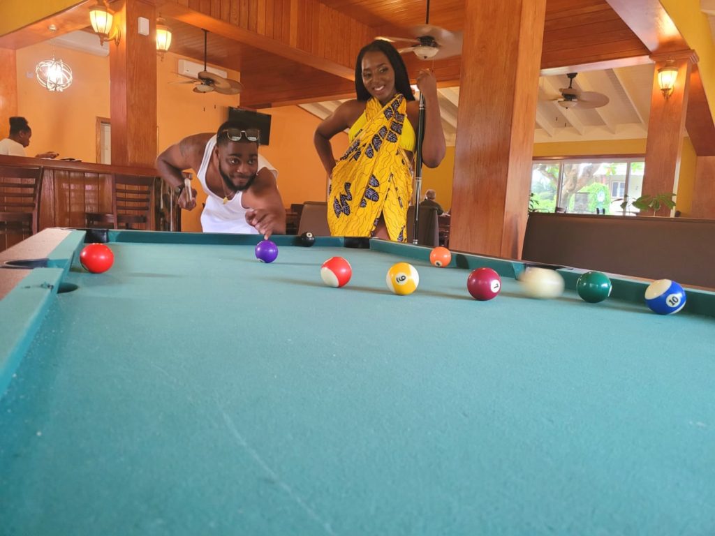 Black girl and caribbean boy playing pool, wearing yellow silk ankara Ngaska print at at 
luxury caribbean resort, Dominica