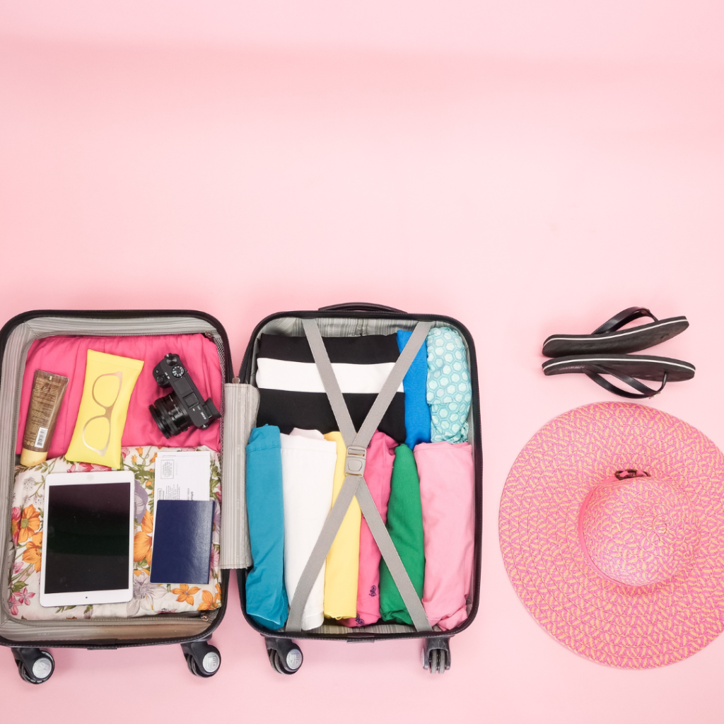 How to Pack Your Suitcase to Maximize Space - suitcase being packed with a camera, a tablet, some clothes, and a pink vacation hat and black slippers, all in a pink photo background.