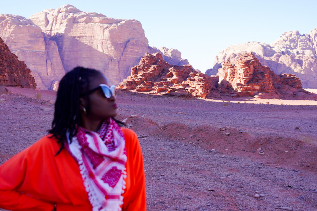 What to wear in Jordan - Travel with Clem, beautiful black influencer wearing an orange flowy dress with a red and white bedouin scarf in Wadi Rum