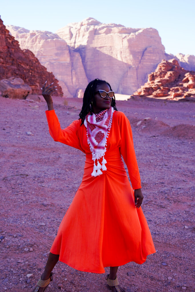What to wear in Jordan - Travel with Clem, beautiful black influencer wearing an orange flowy dress with a red and white bedouin scarf in Wadi Rum