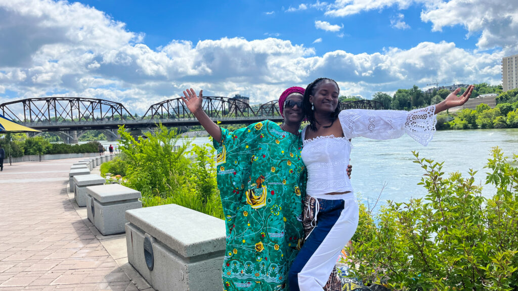 Travel with Clem and her mom posing in beautiful African inspired clothing from Ngaska and standing in front of the South Saskatchewan river - Things to do in Saskatoon